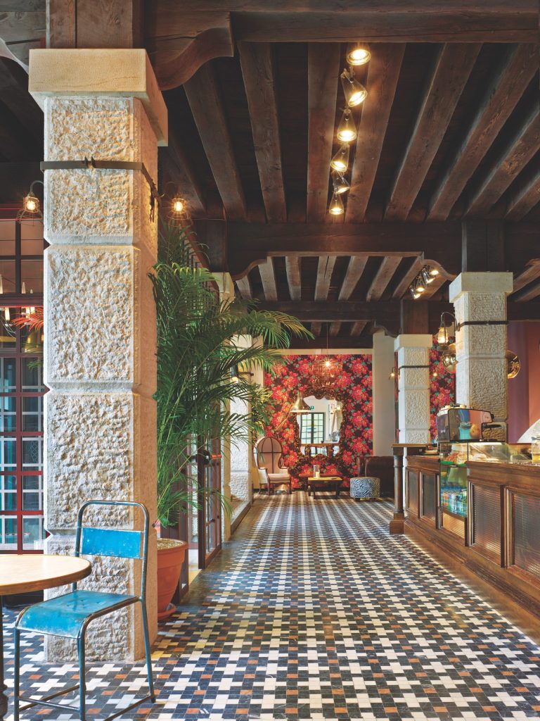 A hallway in a hostel with tiled floors that lead to a seating space with red floral wallpaper behind it. 