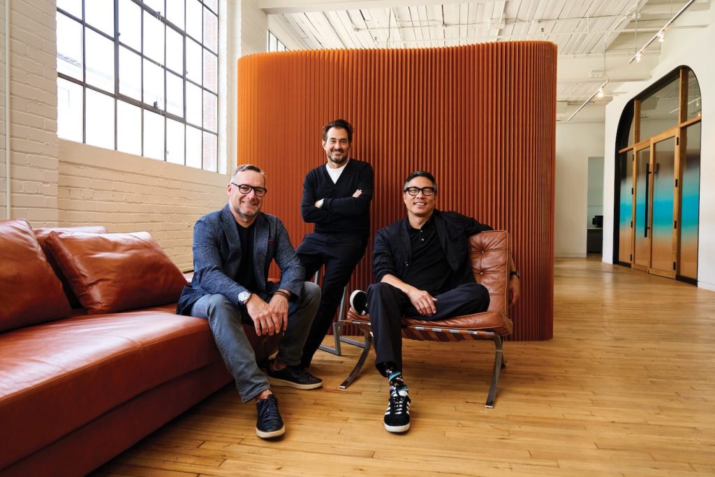 Three men sit on red chairs in a bright studio office space.