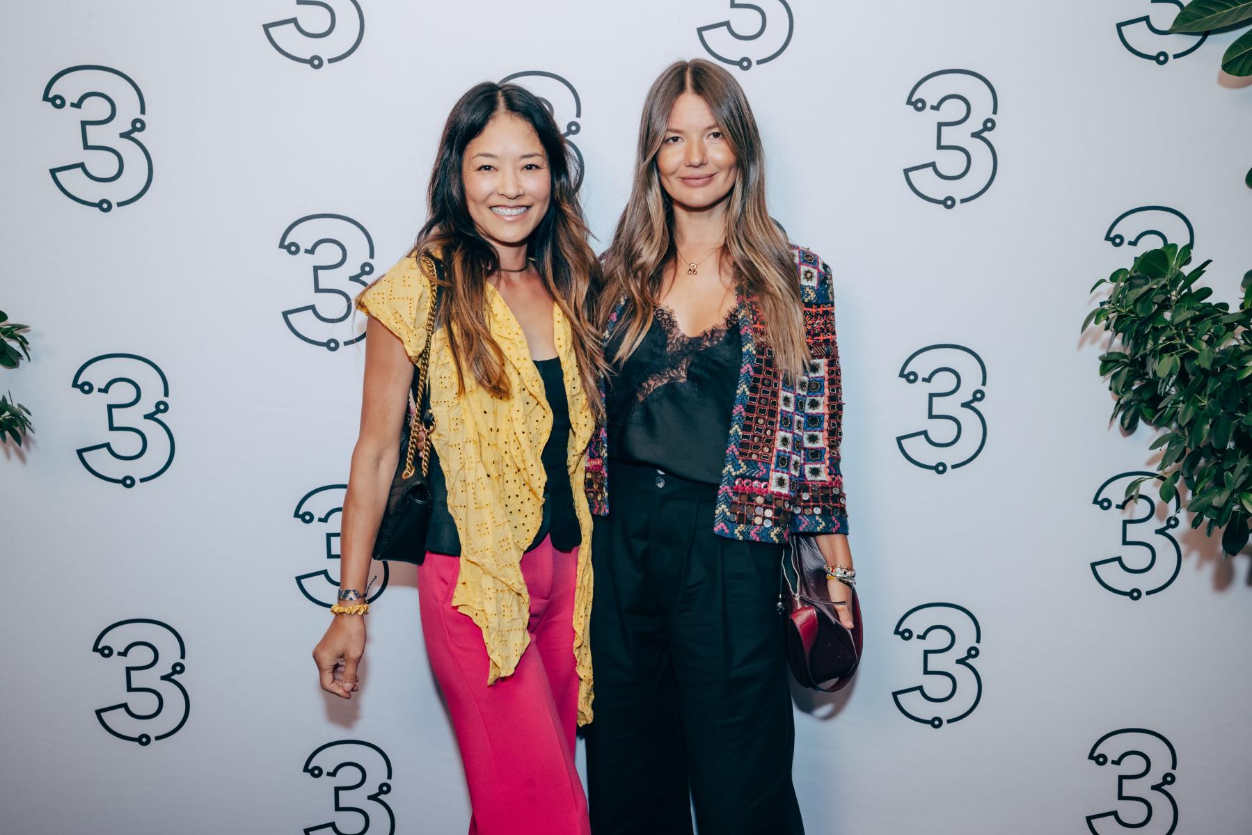Two women posing together at a celebration for Three Magazine's first anniversary, with a backdrop featuring the magazine's logo and greenery, highlighting cultural connection and community.