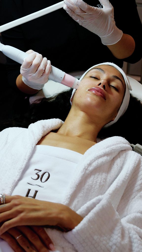 Woman lies down wearing a white spa robe while an aesthetician gives her a facial treatment. 
