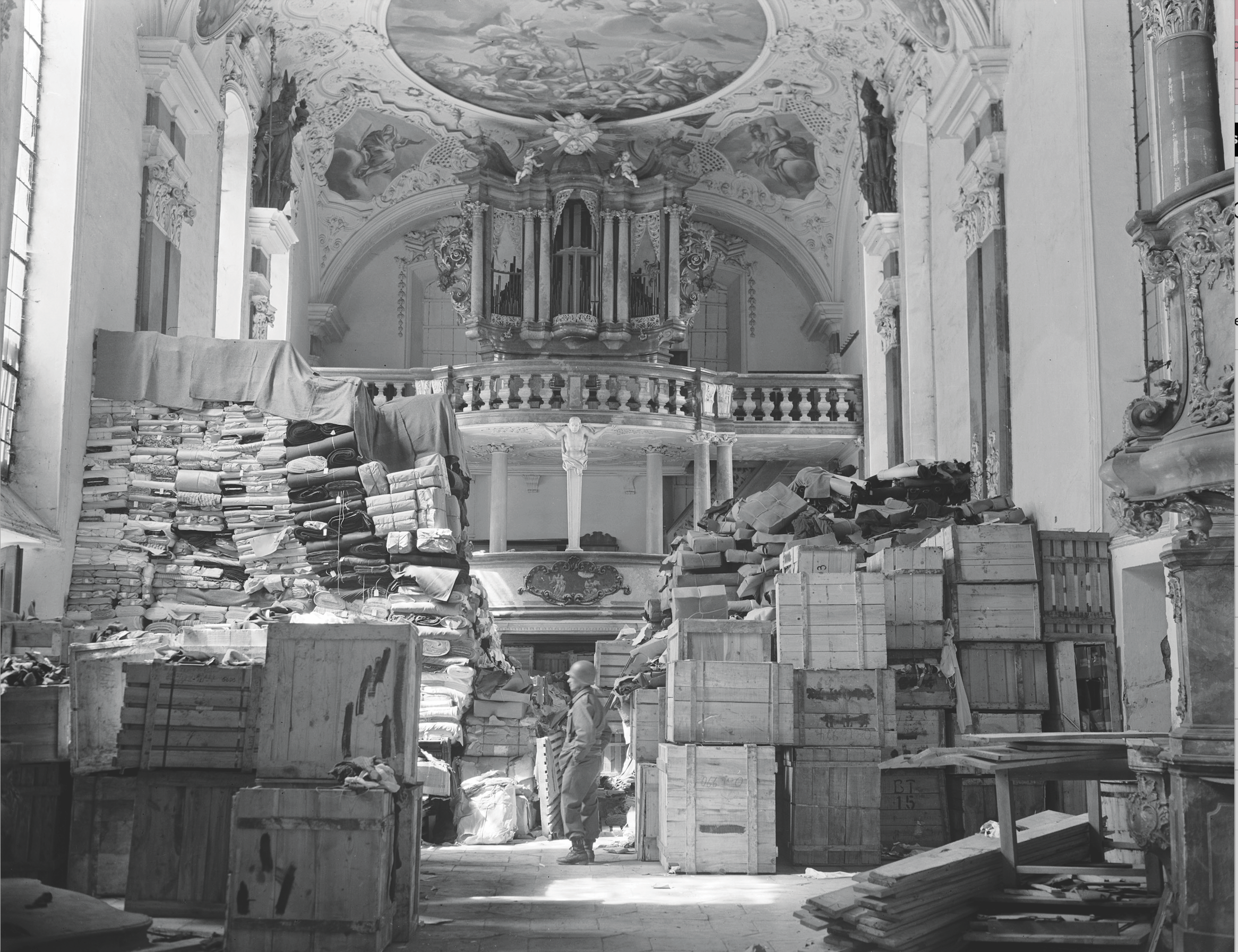 Interior of a grand hall with ornate architecture, featuring stacks of crates and textiles, highlighting the preservation of cultural artifacts.
