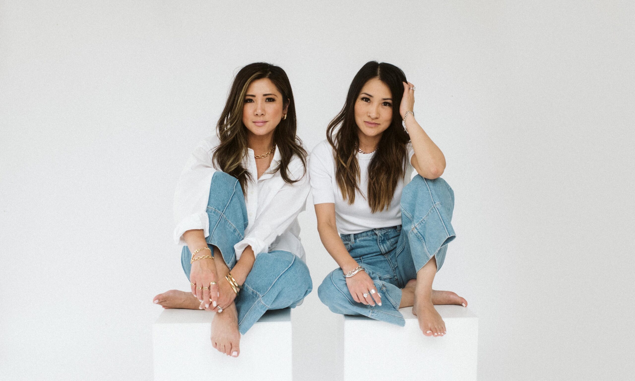 Two women seated on white pedestals, wearing casual outfits with denim jeans and stylish jewelry, representing a contemporary fashion aesthetic related to the jewellery design industry.
