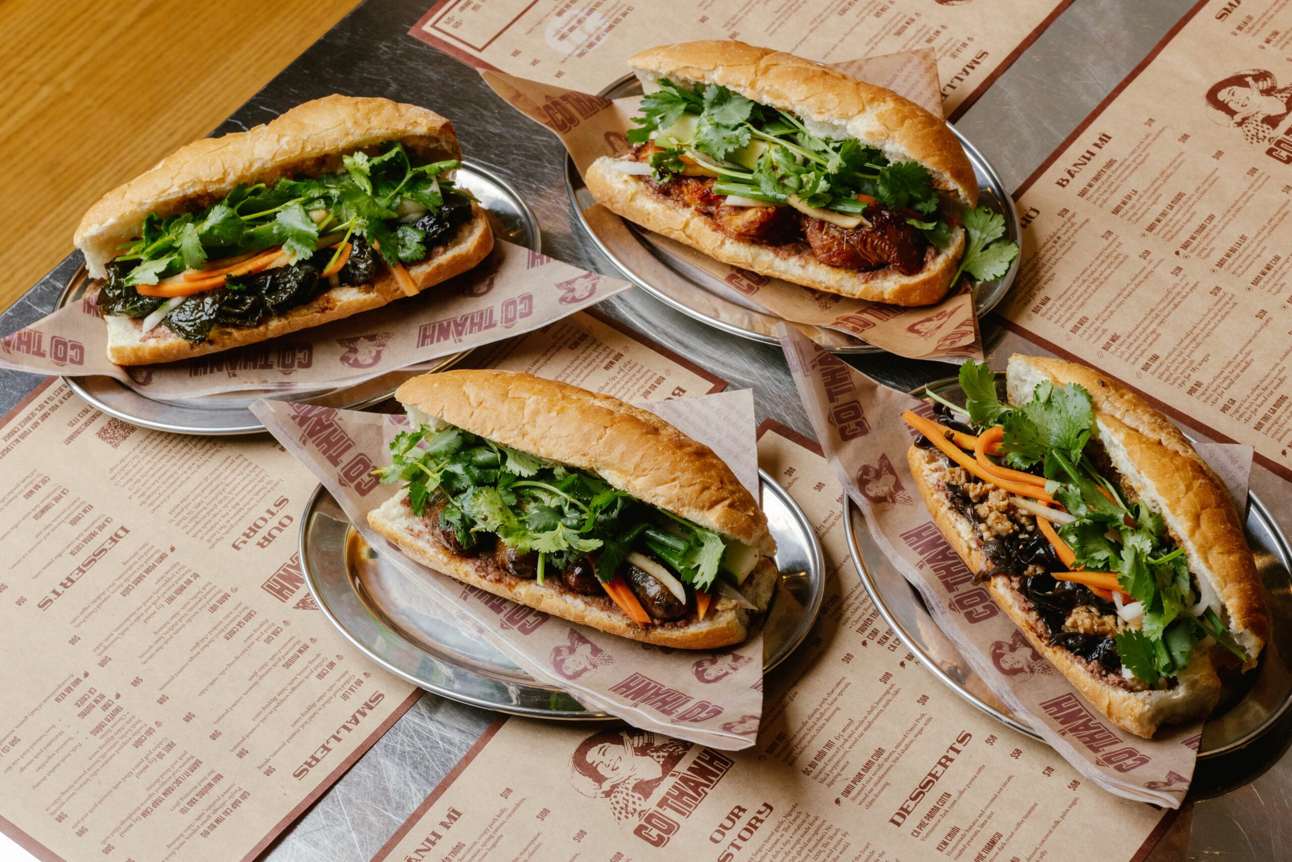 B&aacute;nh m&igrave; sandwiches filled with fresh herbs, vegetables, and meats on metal trays, with a menu backdrop showcasing the global evolution of this iconic dish.