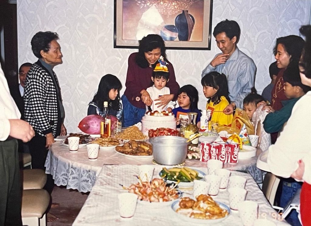Evelyn’s family stands around a dining table full of food as they prepare to cut a birthday cake