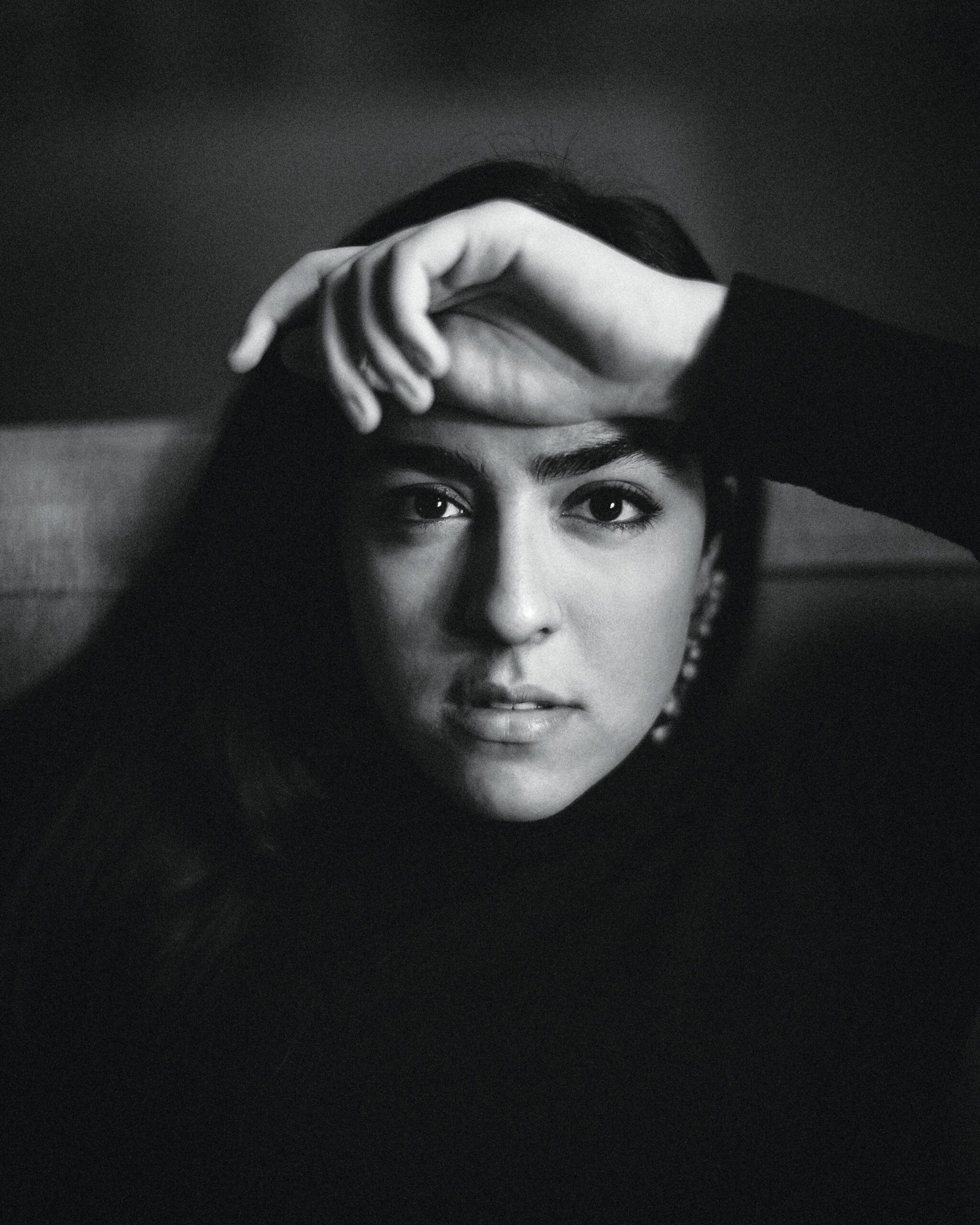 Portrait of fashion designer Golnar Ahmadian, showcasing her expressive gaze and hand resting on her forehead, in a black and white aesthetic, reflecting the artistic depth of her work in the fashion industry.