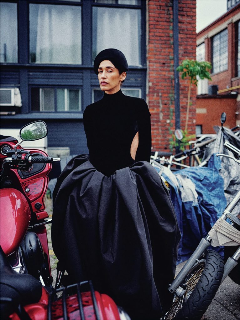 Woman wearing a structured all black outfit stands in front of brick buildings, surrounded by motorcycles.