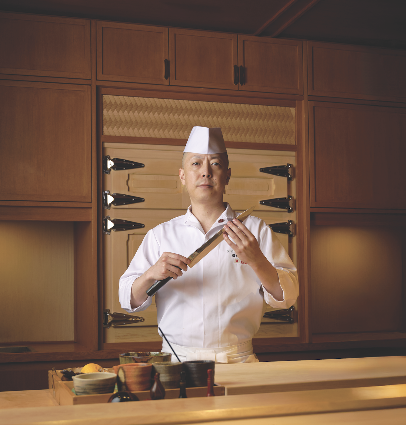 Chef Masaki Saito holding a sushi knife in a traditional kitchen setting, surrounded by ingredients and tools, reflecting culinary identity and craftsmanship.