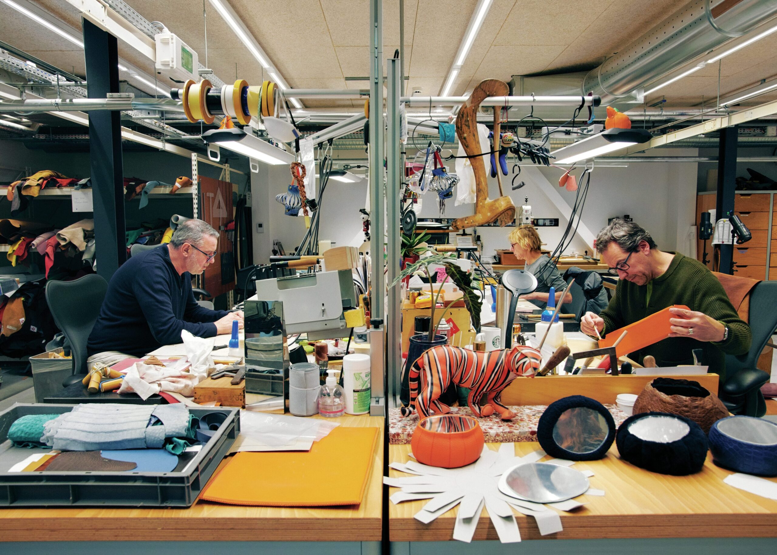 Artisans crafting luxury leather goods in a workshop, featuring colorful materials, tools, and a playful striped tiger figure, reflecting the meticulous craftsmanship of high fashion.