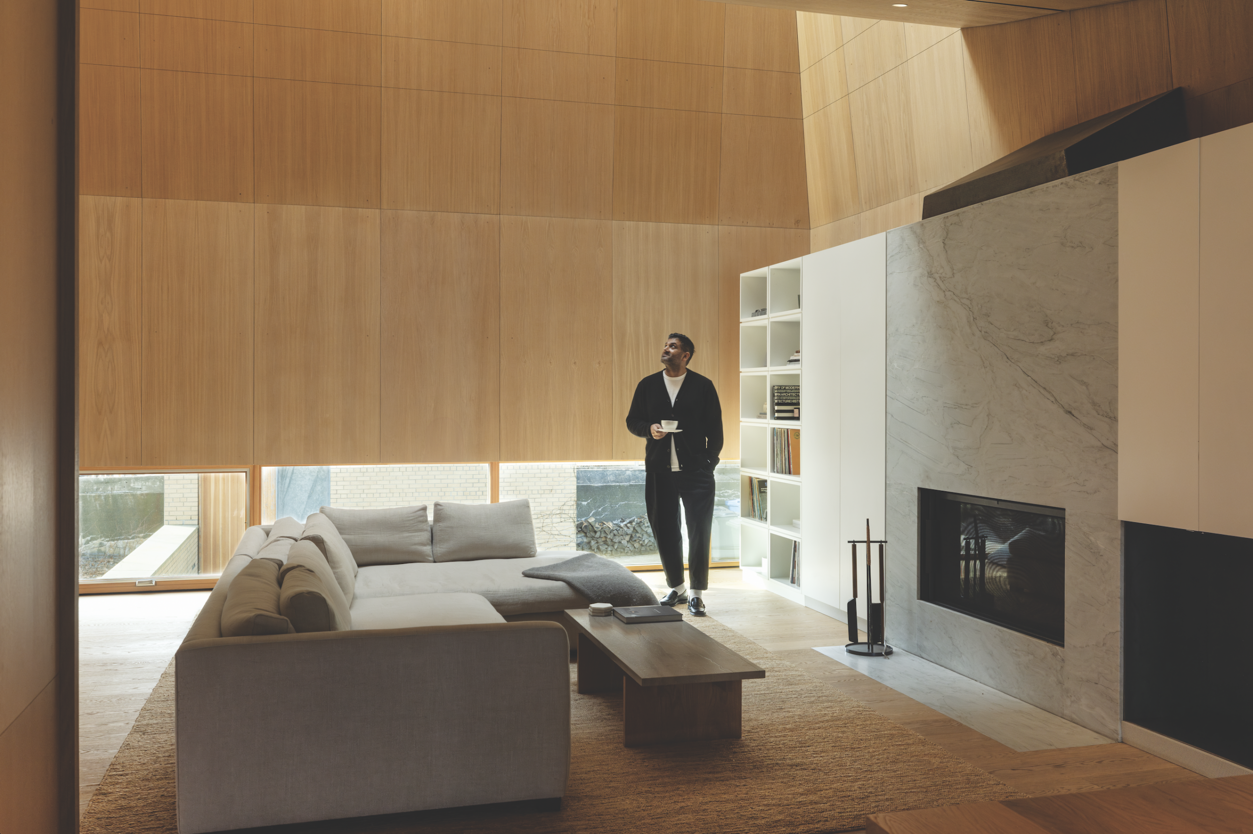 Light-filled interior of a modern home designed by architect Omar Gandhi, featuring a man holding a cup, minimalist furniture, wooden paneling, and large windows.