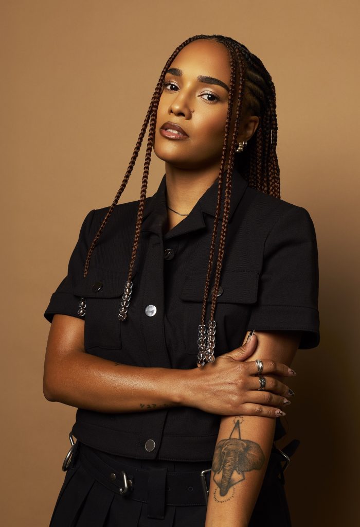 KARABA poses against a tan backdrop wearing a black jumpsuit.