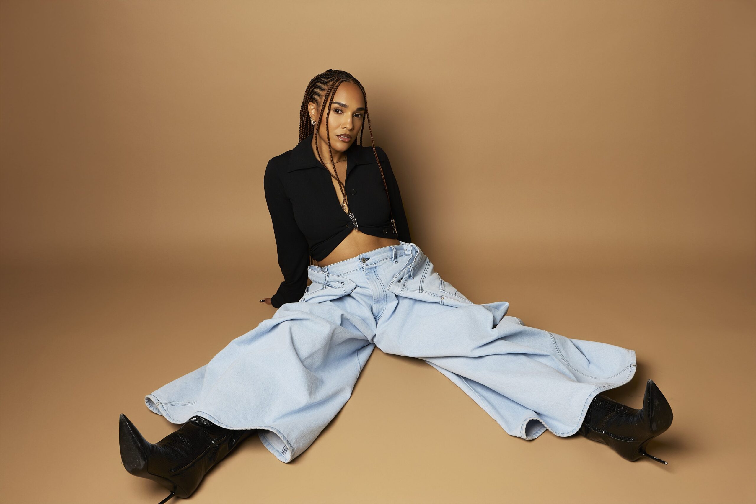 Woman in oversized denim pants and black top, seated against a neutral background, showcasing a stylish and relaxed fashion aesthetic relevant to travel and lifestyle themes.
