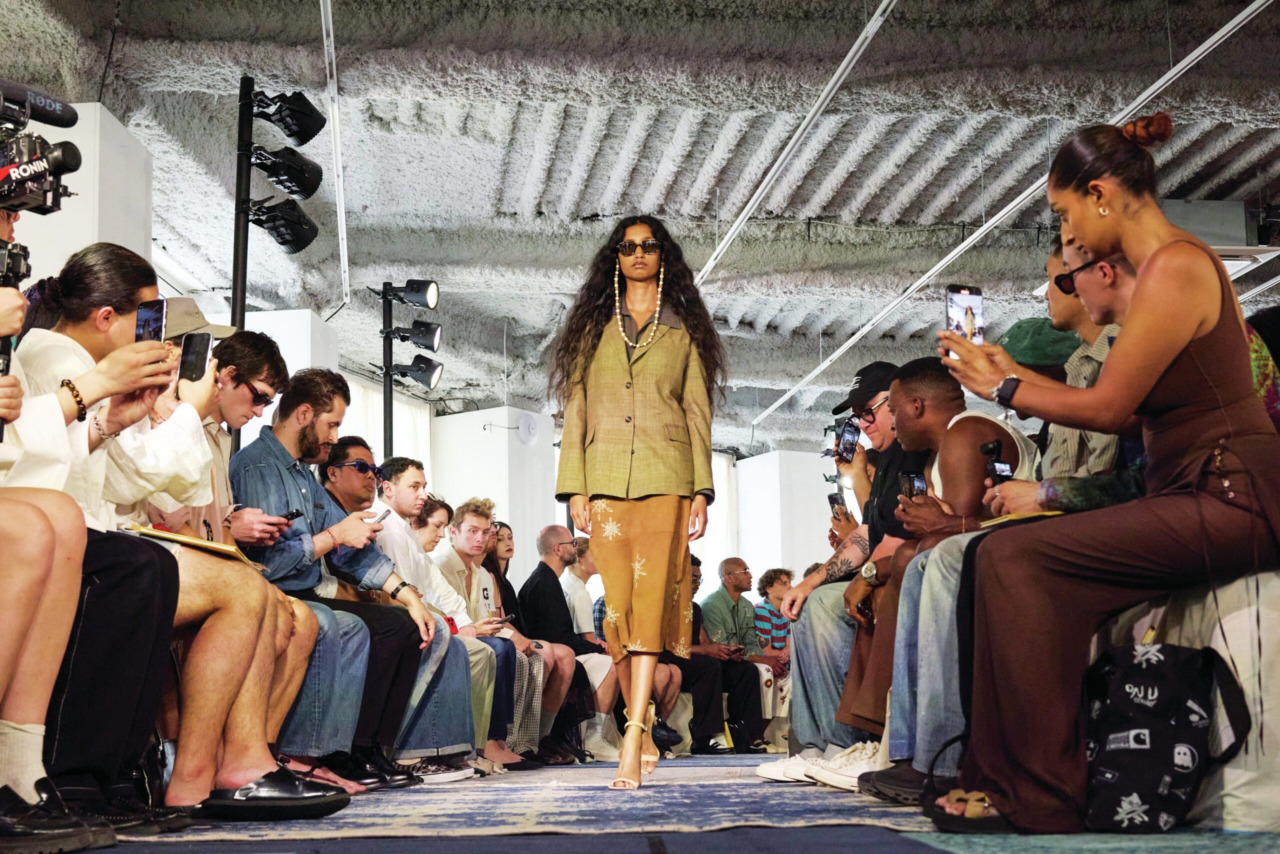 Model walking the runway in a stylish outfit during a menswear fashion show, surrounded by an audience capturing the moment on their smartphones.