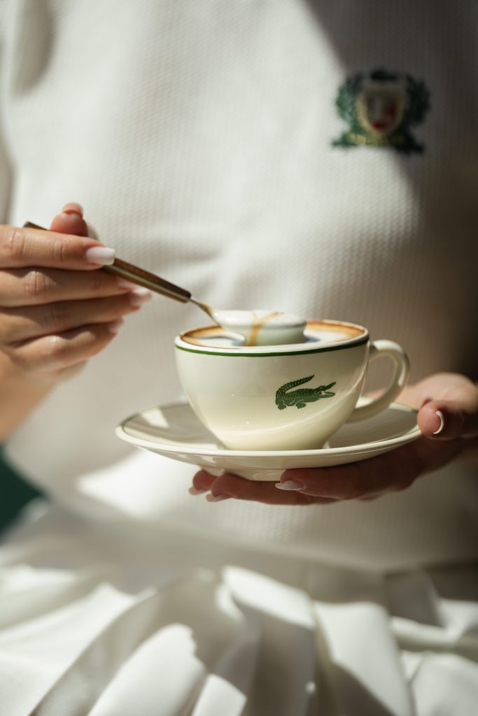 Closeup of a woman holding a cappuccino cup on a small plate. The cup has a dark green crocodile logo on it. 