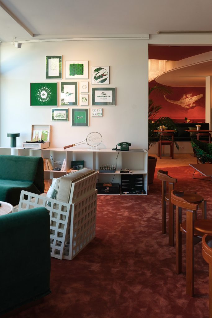 A stylish caf&eacute; interior with green velvet seating, framed artwork on a white wall, retro d&eacute;cor, and warm red carpeting leading to a cozy dining area.