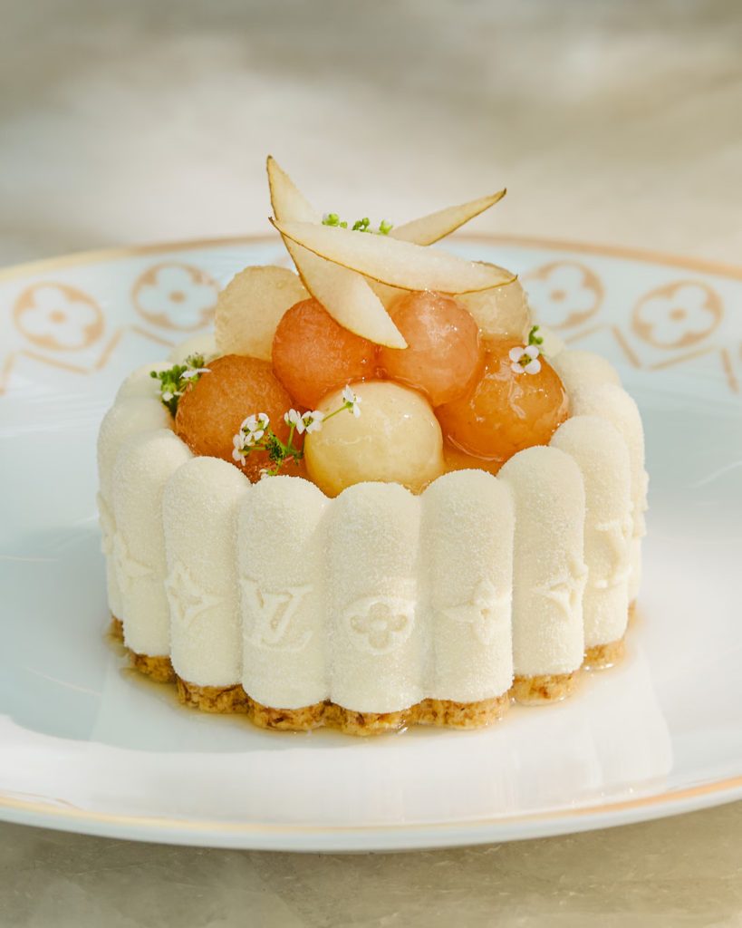 A white mousse cake with embossed Louis Vuitton logos, topped with melon balls, pear slices, and tiny edible flowers. 