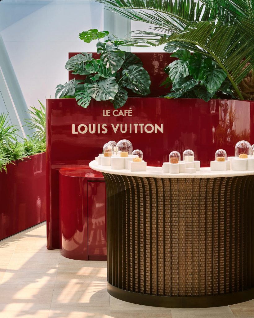 Entrance to Le Café Louis Vuitton featuring a glossy red sign, tropical plants and a counter displaying confections.