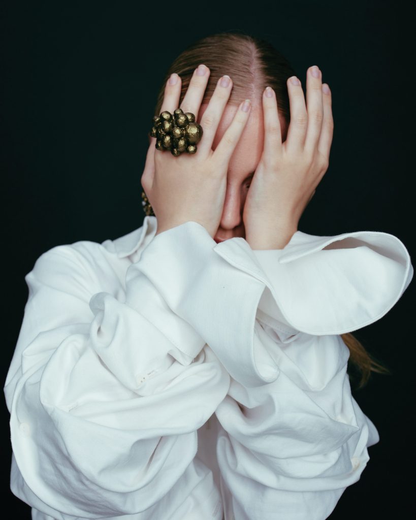 Woman wearing a white button down shirt covers her face with her hands to display a chunky gold ring.