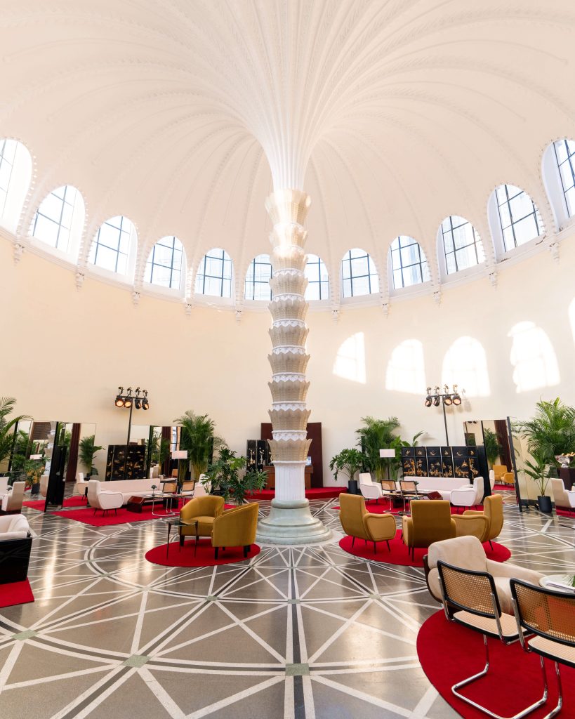 Domed interior lounge with a tall central column, geometric patterned floor, red rugs, and modern seating arranged among indoor plants.