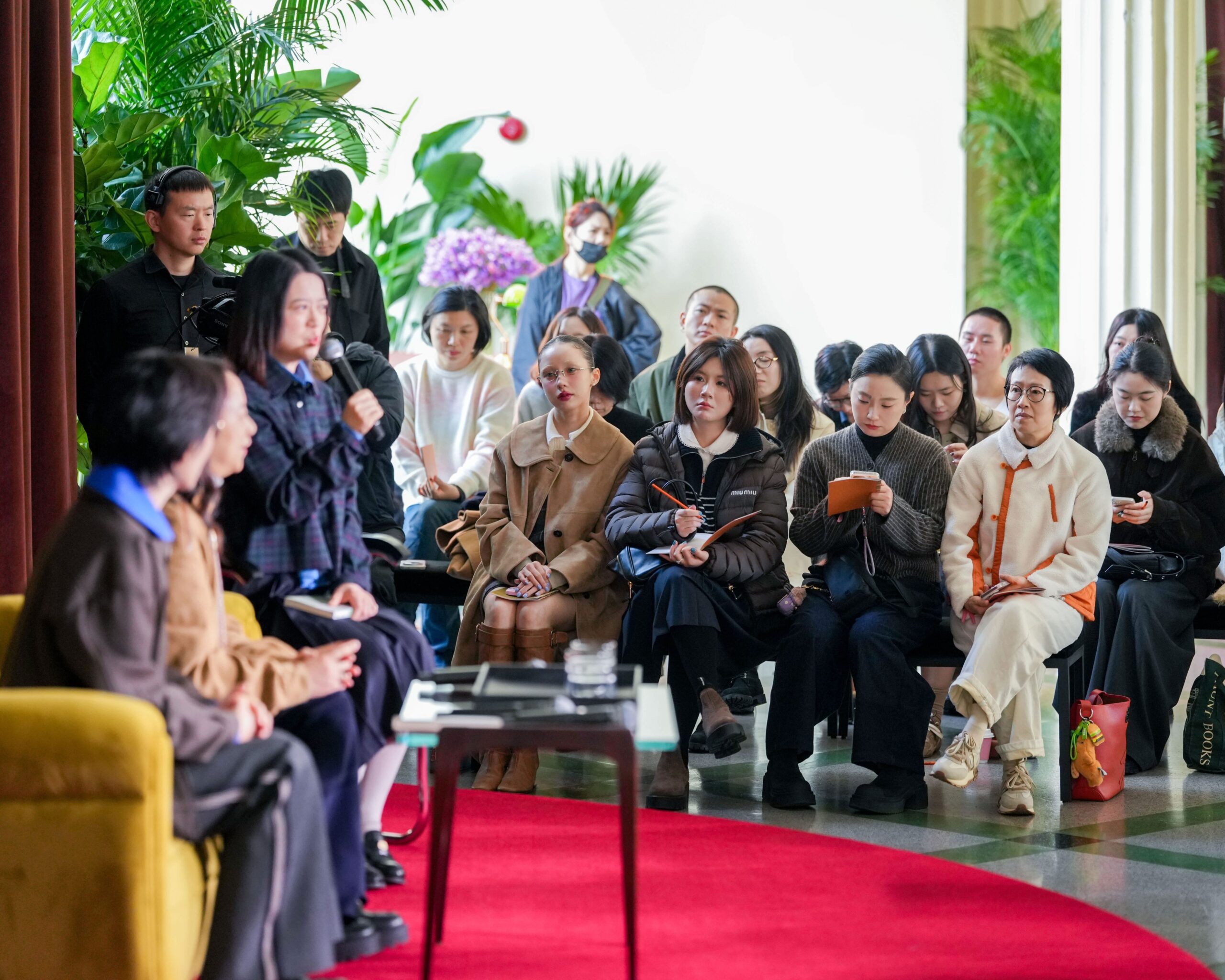Audience engaged in a literary discussion at Miu Miu's event in Shanghai, featuring stylish attendees and a speaker with a microphone, surrounded by lush greenery.
