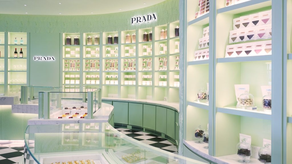 A pastel-green Prada caf&eacute; interior with curved glass display counters and backlit shelves filled with jars, boxes, and packaged sweets.
