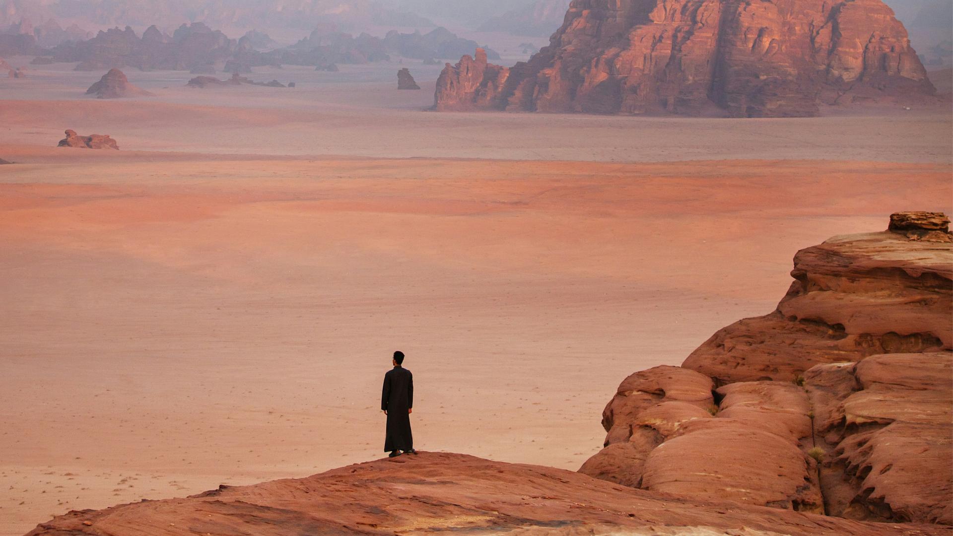 Person in traditional attire standing on a rocky outcrop overlooking a vast desert landscape at sunset, symbolizing Saudi Arabia's vision for future urban development.