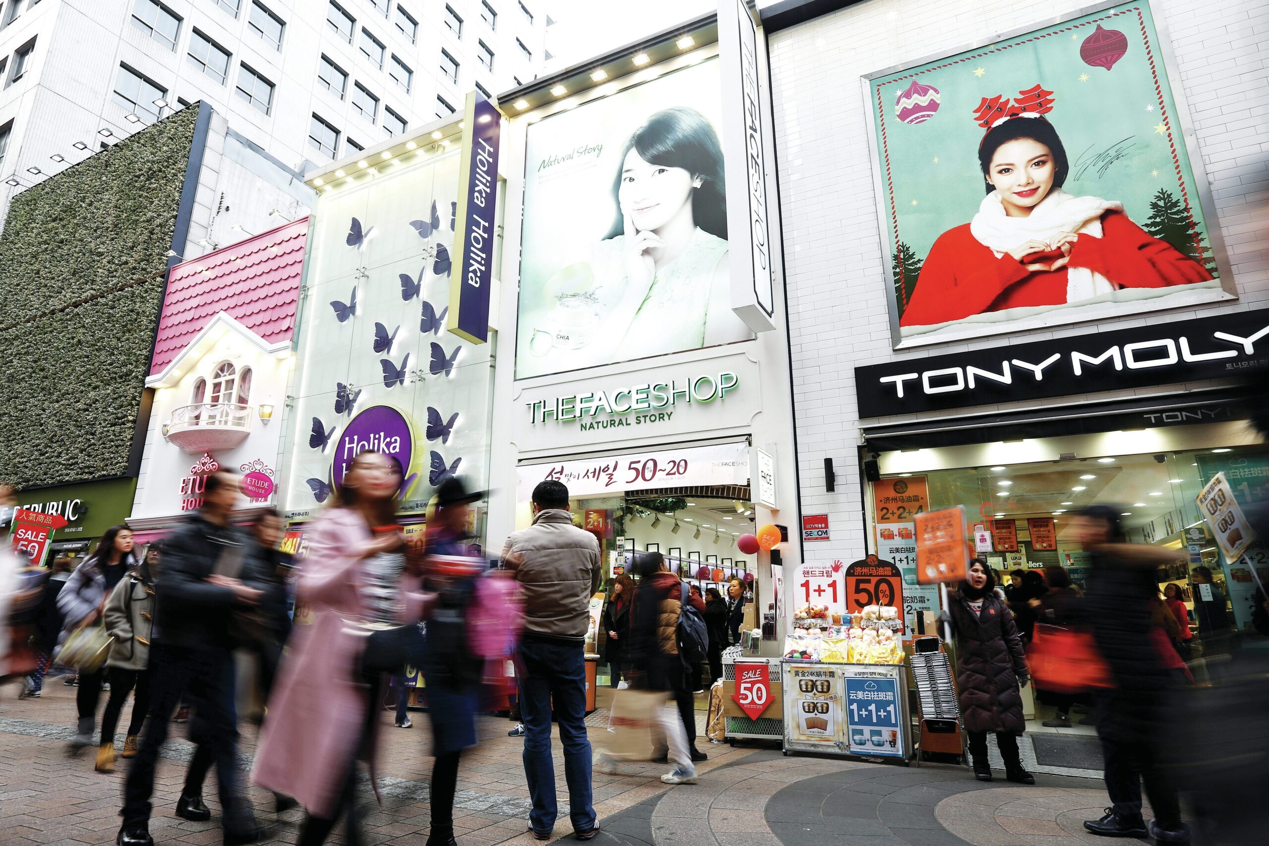 Crowded beauty district in Seoul featuring storefronts of The Face Shop and Tony Moly, with vibrant advertisements and shoppers exploring K-Beauty products.