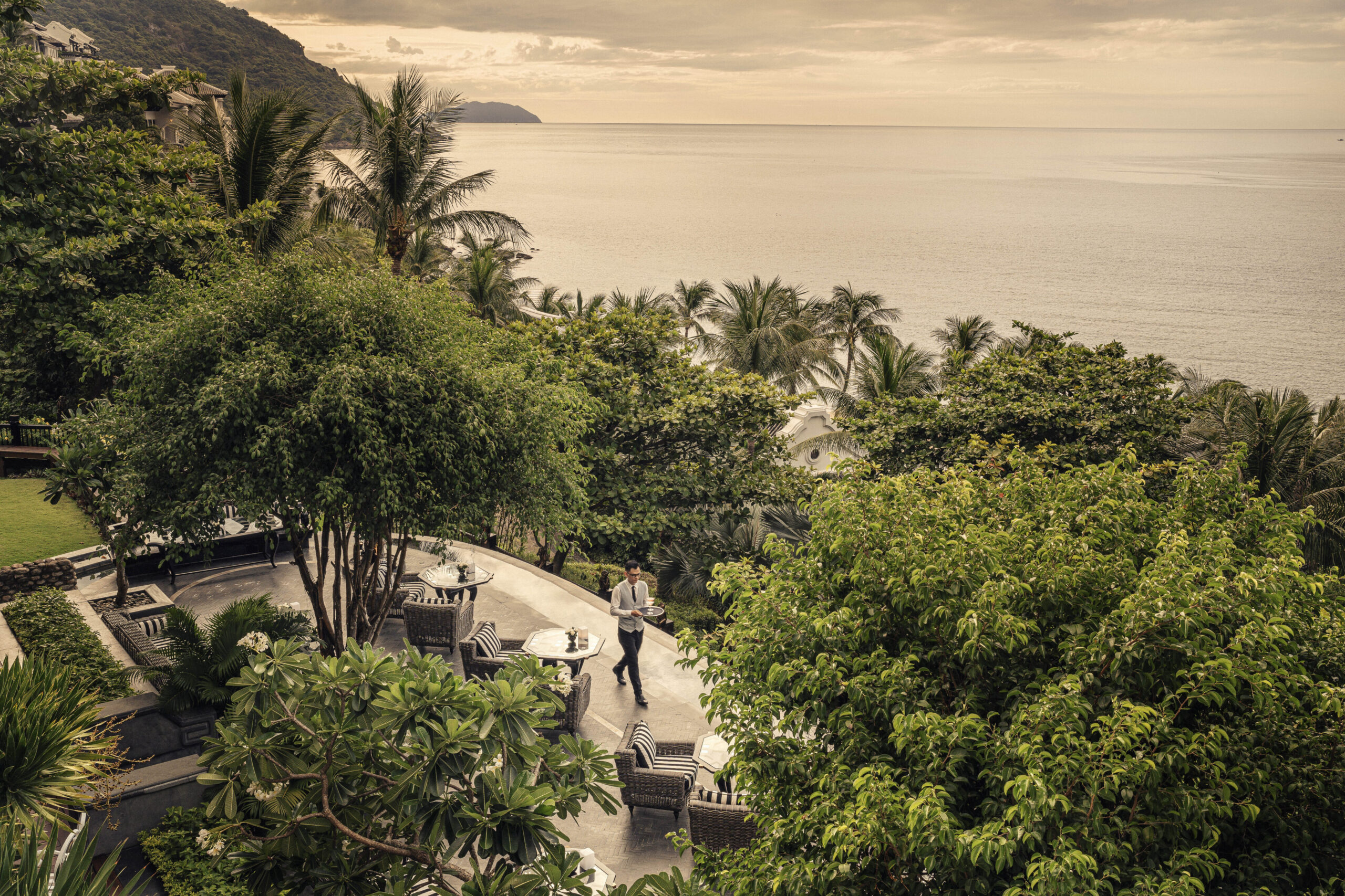 Luxurious outdoor dining area overlooking the ocean, with a waiter serving guests amidst lush greenery and tropical palm trees in a serene setting.