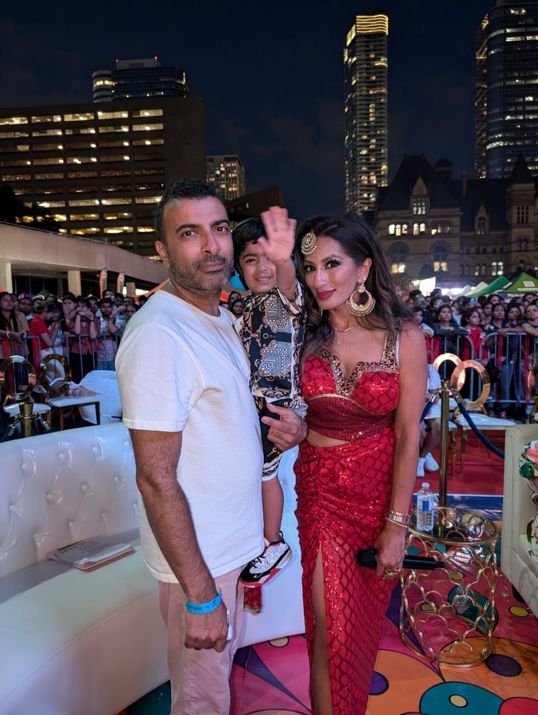 Uppekha Allani poses with her husband and son at an event in Toronto's Nathan Phillips Square. Allani is wearing a red sparkly Indian dress. 