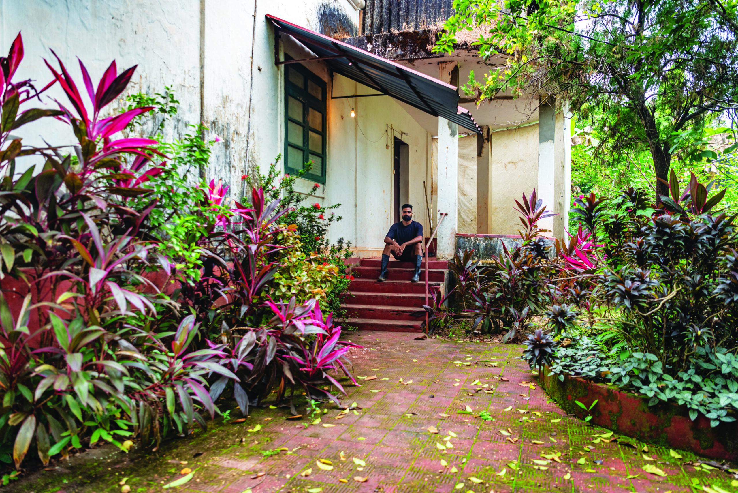 Man sitting on steps of a house surrounded by vibrant plants and greenery, reflecting themes of identity and cultural connection.