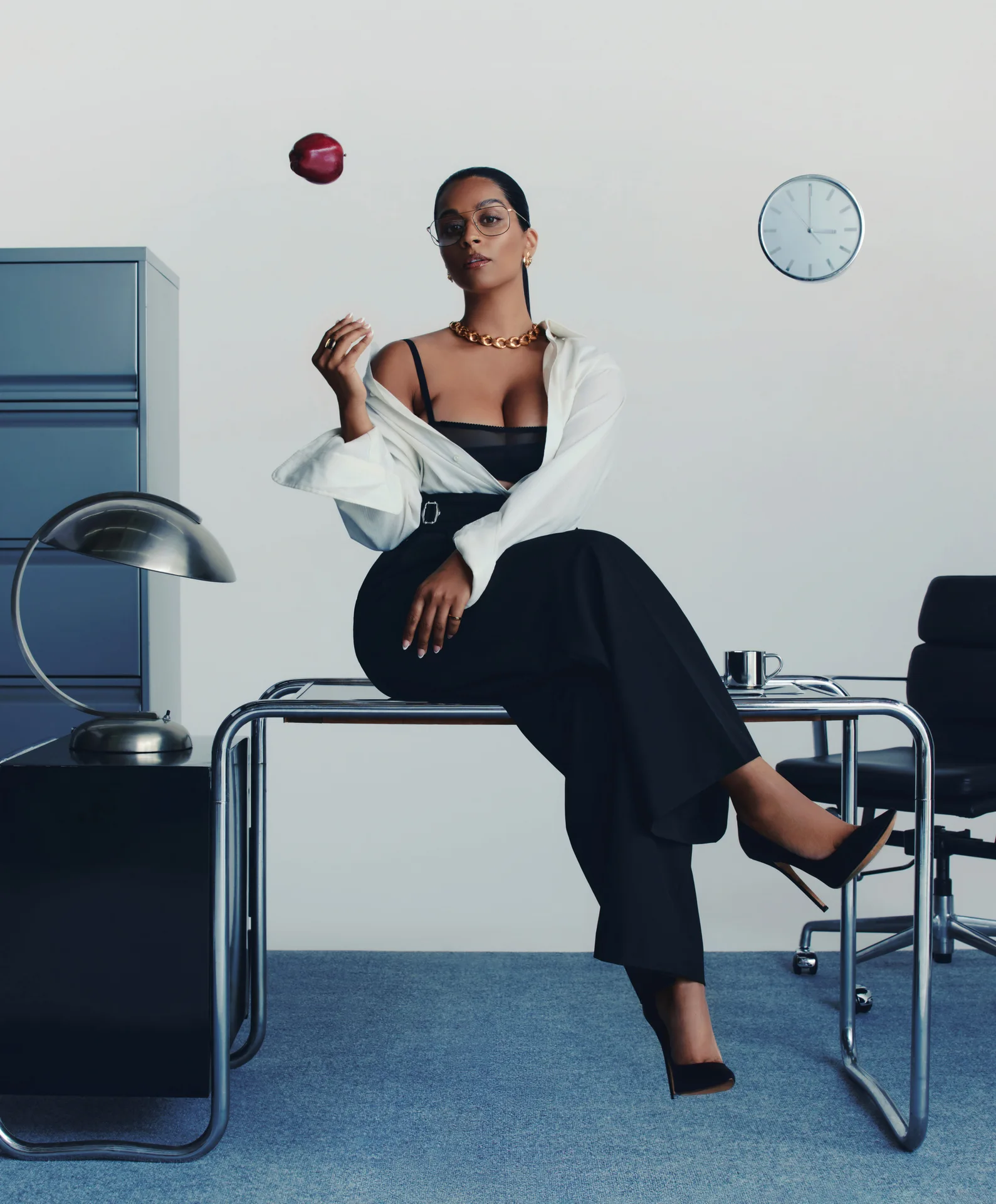 Lilly Singh sitting confidently on a desk in a stylish outfit, holding an apple, with modern office elements like a clock and lamp in the background, reflecting themes of empowerment and breaking taboos in her film "Doin' It."