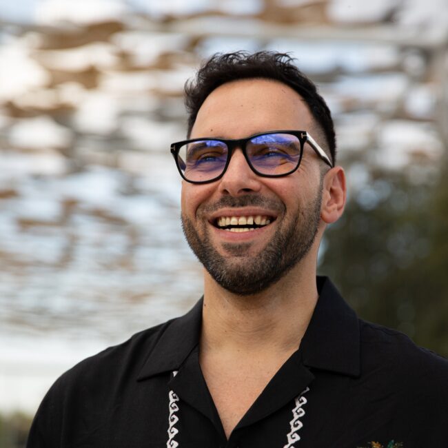 Abdulah Al-Ghoul smiling outdoors, wearing glasses and a black shirt, reflecting themes of multicultural identity and global storytelling in Three Magazine.