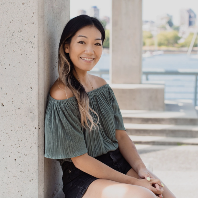 Erica Fong, smiling and seated casually against a concrete pillar, wearing a green off-the-shoulder top and black shorts, with a waterfront view in the background, representing the editorial leadership of Three Magazine.