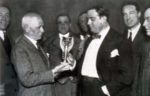 Historic sepia photo of a footballer holding the Jules Rimet Trophy while surrounded by officials and supporters.