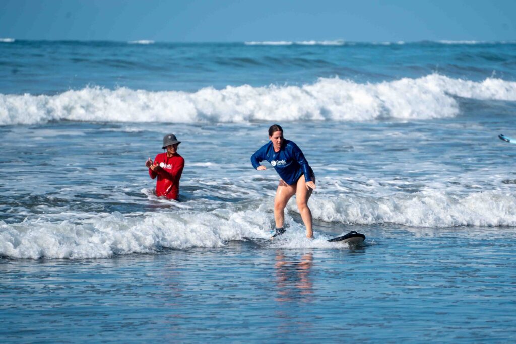 A woman standing on a surf board with a Bodhi Surf + Yoga instructor watching her.