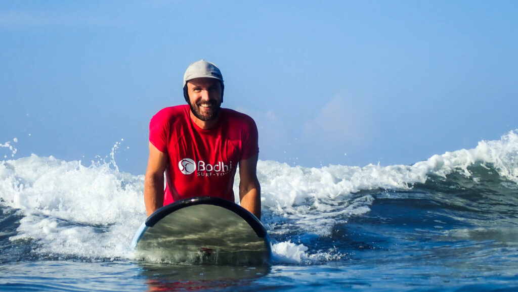 Travis, founder of Bodhi Surf + Yoga, on a surfboard on the beach at Playa Chaman with a warm smile, ready to teach a beginner surf lesson.
