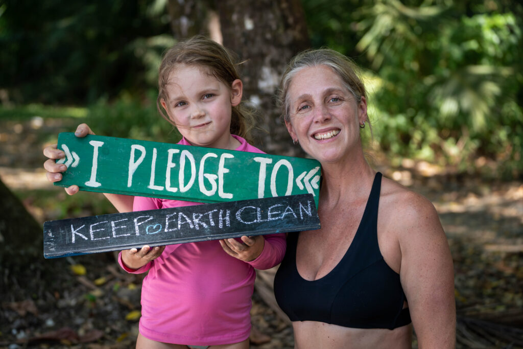 A smiling girl in a pink shirt and a woman hold two hand-painted wooden signs that read "I PLEDGE TO" and "KEEP EARTH CLEAN," standing in a lush, sunlit outdoor setting.