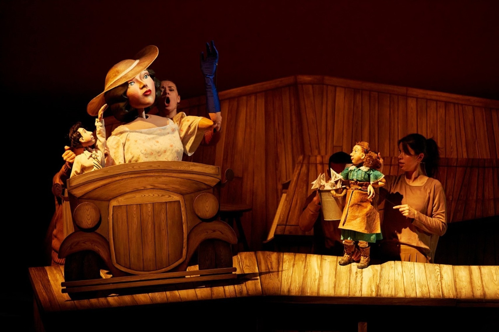Une grande marionnette, portant une robe et un grand chapeau, est dans une voiture avec une autre petite marionnette. À droite, une marionnette de taille moyenne la regarde. Le décor et les accessoires sont en bois.