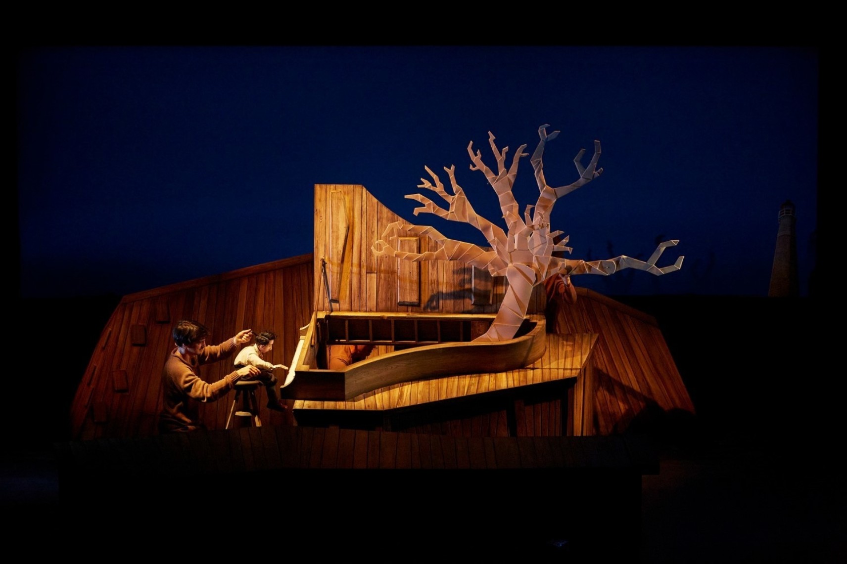 Un marionnettiste manipule une petite marionnette jouant du piano. Un arbre sort du coffre du piano qui est ouvert. Le décor et les accessoires sont faits en bois et le fond est éclairé d'une lumière bleue.