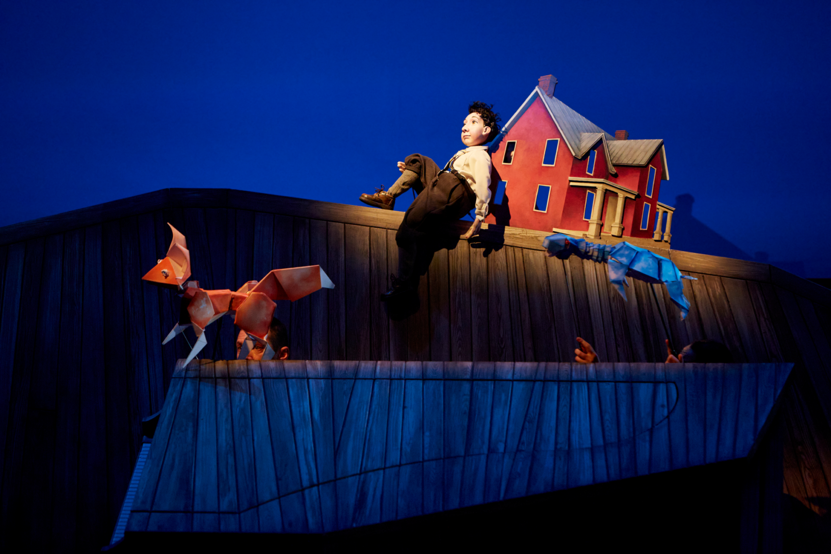 Dans un décor en bois, une marionnette de petit garçon est adossée à une maison rouge. En avant-plan, les marionnettes de renard et de tortue en mouvement. L'arrière-plan est éclairé par une lumière bleue.