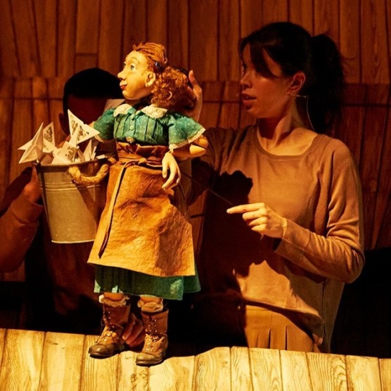Dans un décor en bois, Audrey manipule la marionnette Delphine, une marionnette de taille moyenne portant une robe, un tablier et un sceau plein de papiers.