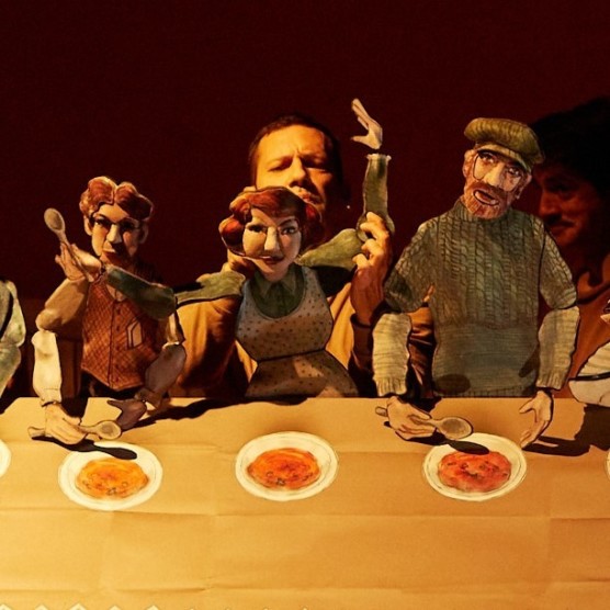 Martin manipule les marionnettes de la Famille de pêcheurs. Trois marionnettes, deux hommes et une femme, sont assises à une table en bois. Devant elles, des assiettes sales.