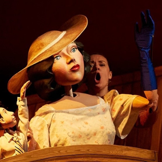 Olena manipule Tante Simone, une grande marionnette en bois portant une robe blanche, du rouge à lèvres et un chapeau à large rebord.