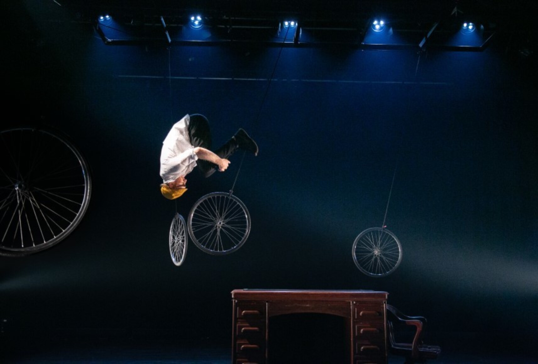 L'interprète fait un salto dans les airs avec une roue de vélo. La scène présente un décor minimaliste avec d'autres roues.