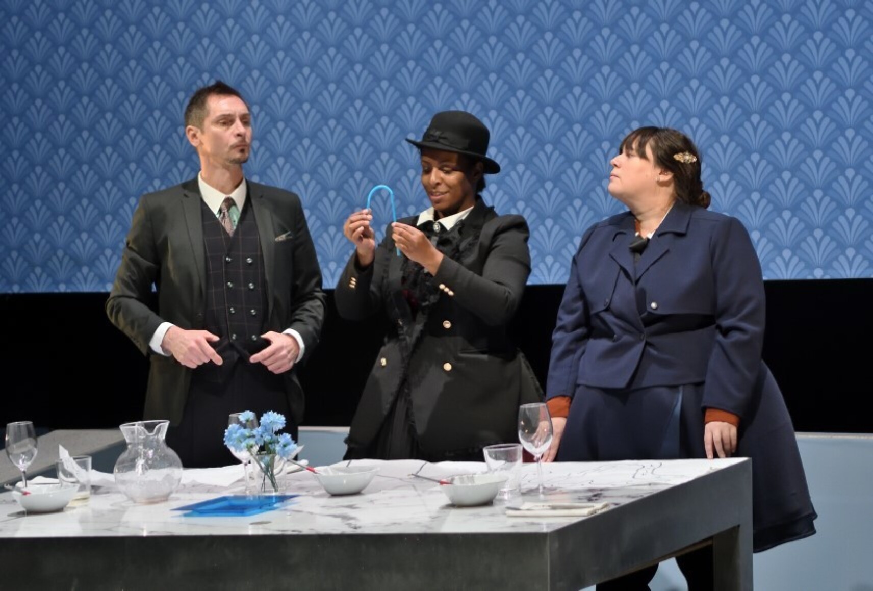 Trois personnages diplomates en tenue officielle se tiennent devant une table. Celle au centre porte un chapeau melon. Leurs costumes sont dans des teintes sombres de gris, noir et marine. L'arrière-plan est un papier peint à motifs art déco bleu.