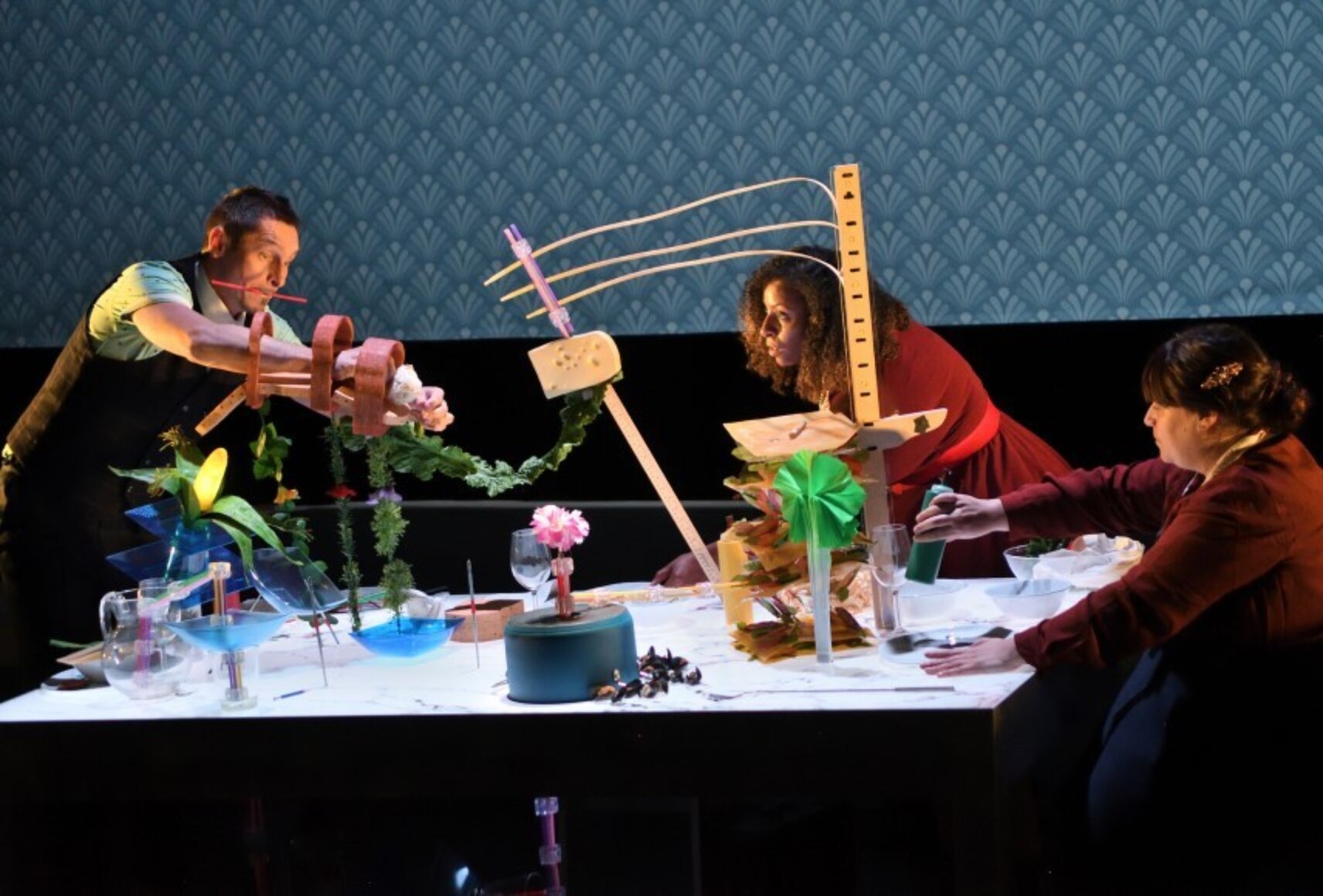 Les trois personnages construisent des sculptures de différente grandeur avec divers aliments. L'arrière plan est un papier peint à motifs bleu.