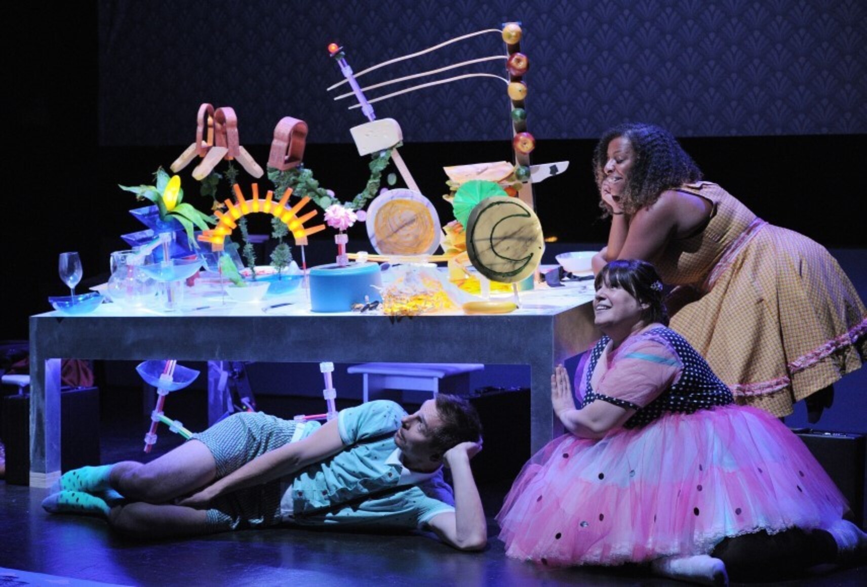 Trois personnages portant des déguisements d'enfants colorés, sont autour d'une table avec diverses sculptures sur le dessus.