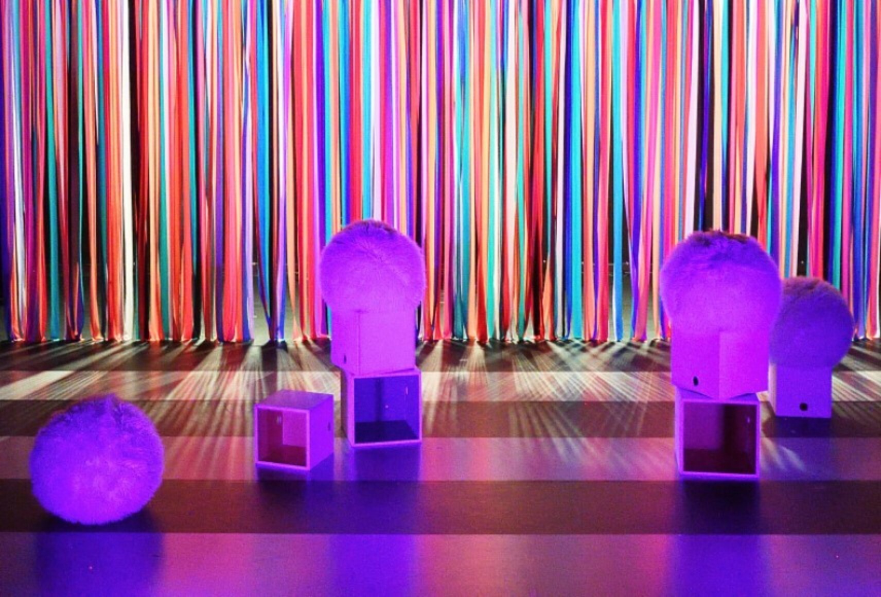 Des cubes et des boules poilues sont sur la scène dans une lumière violette. Au fond, un rideau fait avec des lanières de tissus de toutes les couleurs.