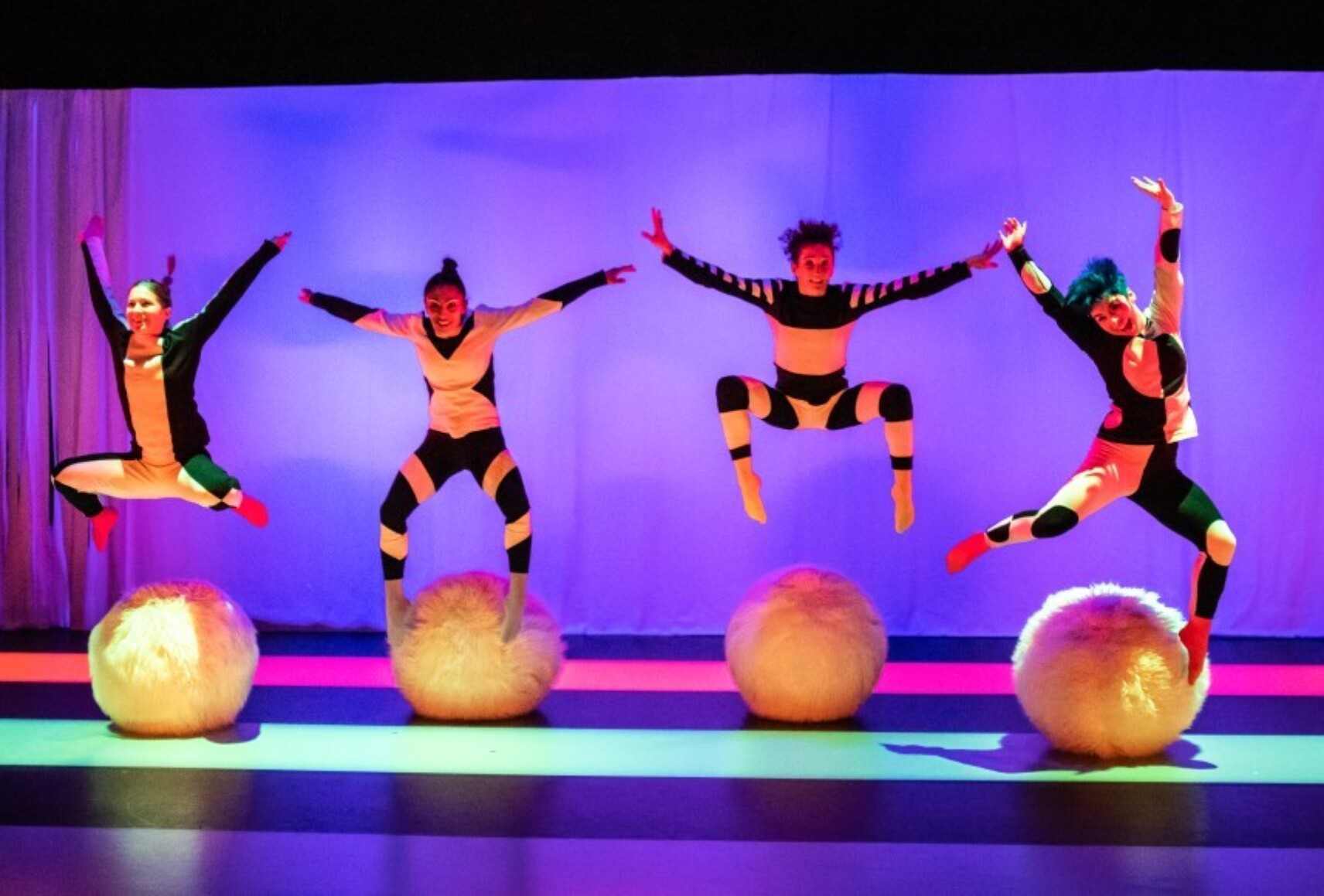 Quatre danseuses sautent au-dessus des boules poilues en faisant de grands gestes. Elles sont habillées en noir et blanc. L'arrière plan est mauve et la lumière trace de larges bandes rose et turquoise au sol.