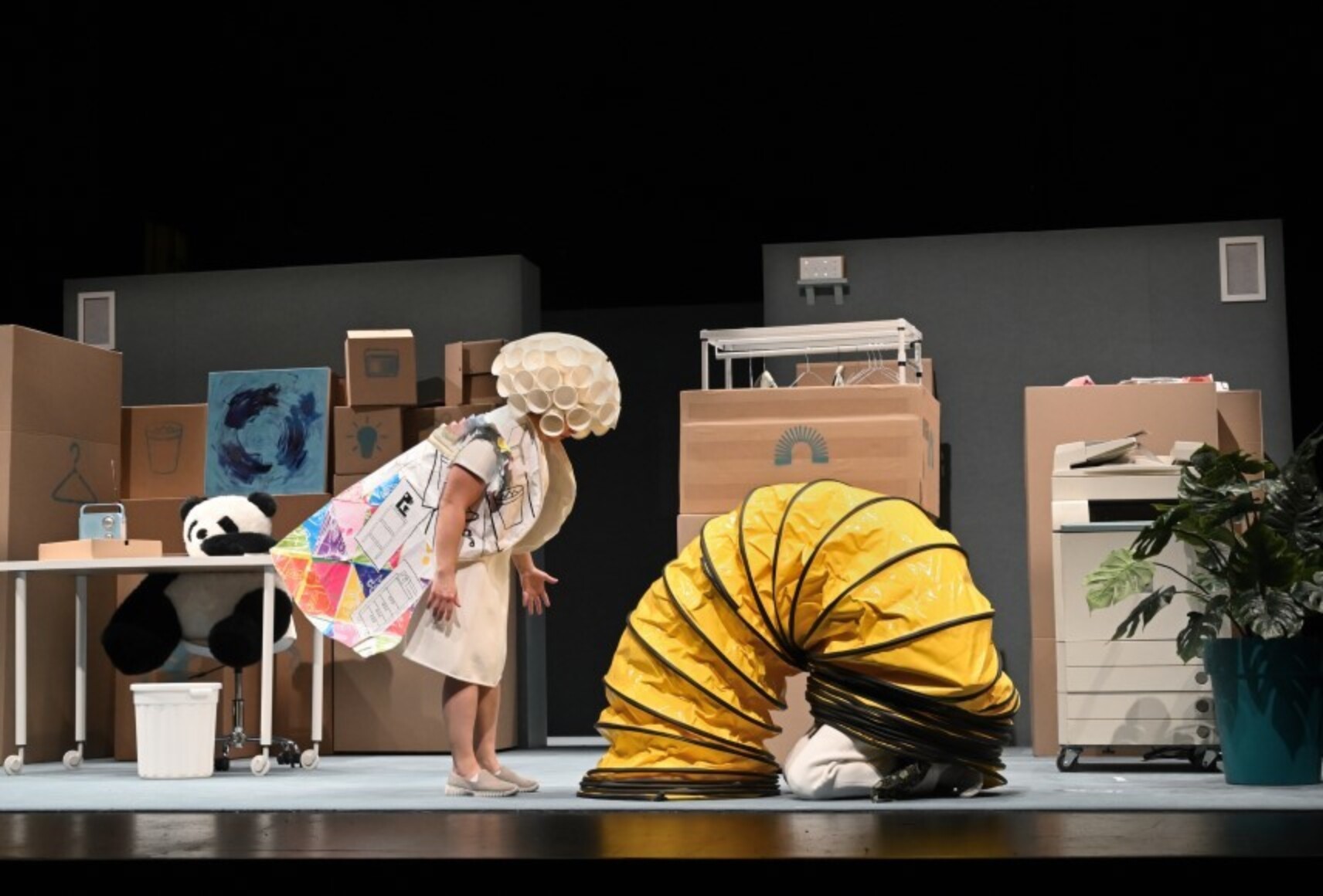 Décor de bureau avec beaucoup de cartons, une radio, une plante, une photocopieuse, une peluche de panda. Attention porte une cape colorée et un chapeau extravagant. Au sol, une sorte de chenille jaune dont ressortent des jambes.
