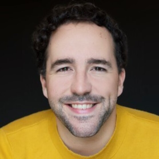 Photo du comédien Jean-François. Il a les cheveux bruns bouclés, une barbe de quelques jours, et un chandail jaune. Il sourit en regardant l'objectif.